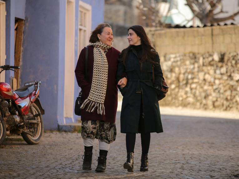 Zwei Frauen gehen nebeneinander eine Straße entlang. Sie haben einander eingehakt und scheinen Mutter und Tochter zu sein.
